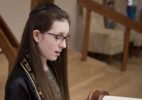 Pictured here as a middle schooler, Mina wears her Tallit (a Jewish prayer shawl ceremonially given to the B’Nai Mitzvah at the start of the ceremony, symbolizing the welcoming of their new status as a Jewish adult) and reads from the Torah (תורה) - Jewish scripture written in Biblical Hebrew. This photo was not taken during her B’Nai Mitzvah ceremony, since phones are not allowed in the sanctuary during the ceremony. Photo courtesy of Mina Koffron.